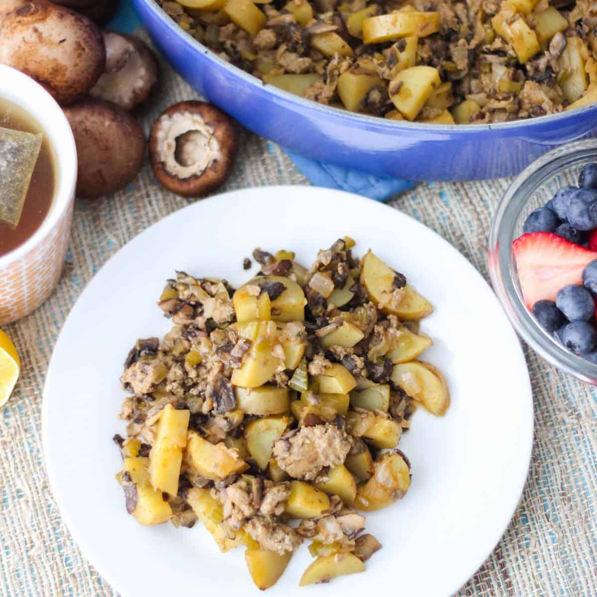 white plate of mushroom hash next to a bowl of berries, cup of tea, scattered mushrooms, and a pot of hash