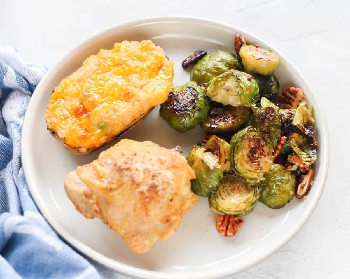 plate with a chicken thigh, roasted Brussels sprouts and pecans, twice baked potato next to a blue napkin