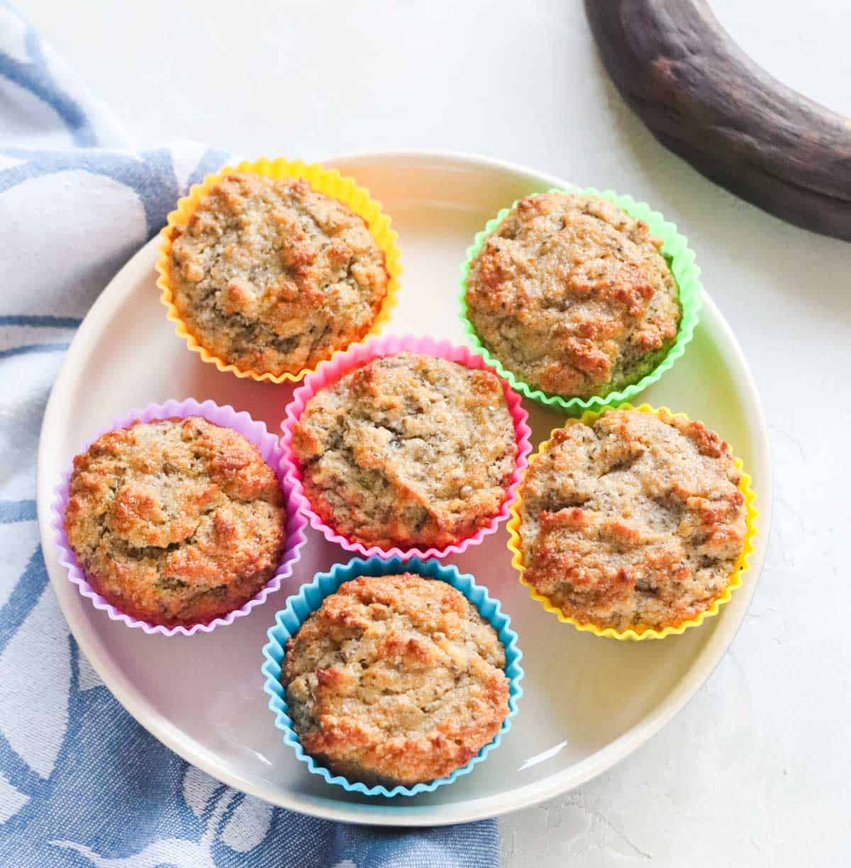 six muffins in colorful silicone muffin liners on a beige plate next to an old banana and blue napkin