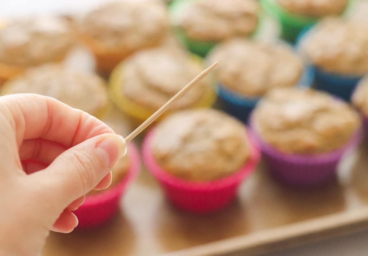 hand holding clean toothpick with baked muffins on baking sheet