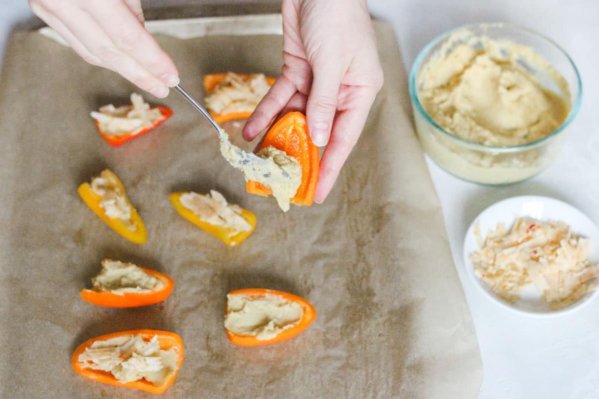 parchment-lined baking sheet with hummus filled mini peppers and a hand spooning hummus onto a pepper