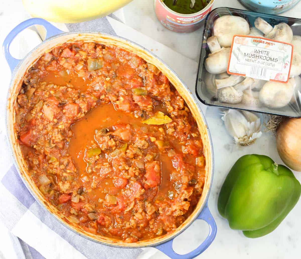 pot of turkey meat sauce with tomatoes next to green bell pepper, head of garlic, onion, and container of mushrooms