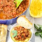 bowl of turkey meat sauce with a spaghetti squash next to garlic bread, fresh basil, and a bowl of prepared spaghetti squash with meat sauce on top