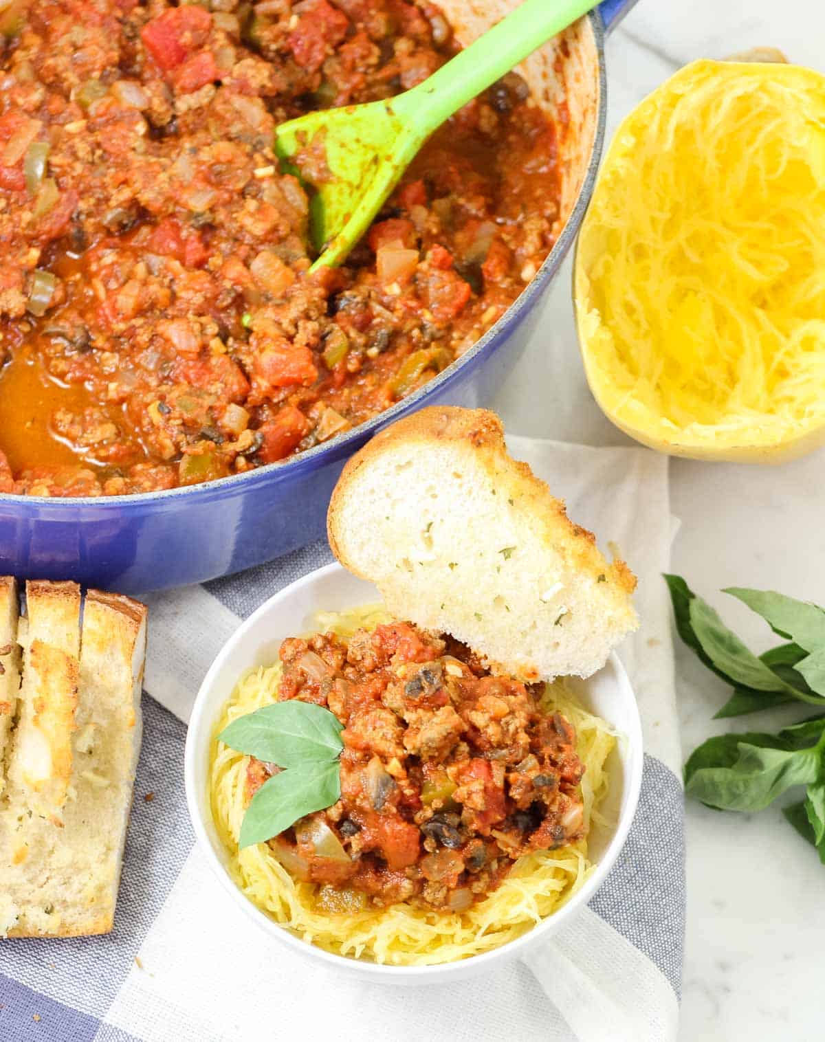 bowl of turkey meat sauce with a spaghetti squash next to garlic bread, fresh basil, and a bowl of prepared spaghetti squash with meat sauce on top