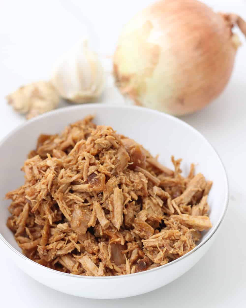 white bowl with shredded pork next to yellow onion, fresh garlic, fresh ginger.