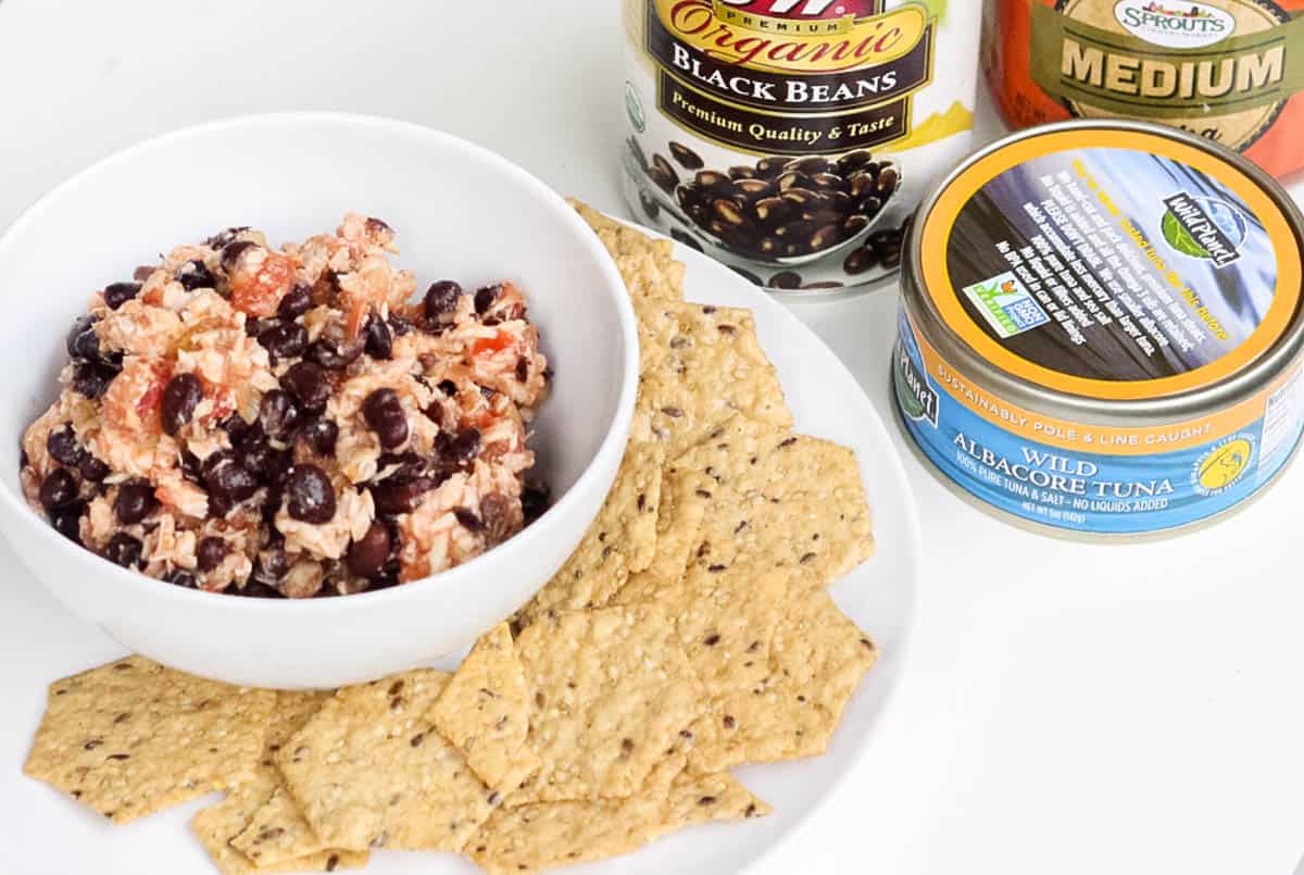 crackers on a white plate with a white bowl with salsa tuna salad, can of black beans, can of tuna, and jar of salsa.