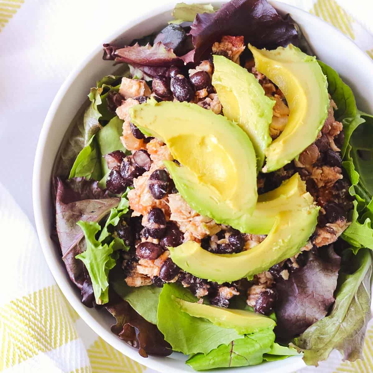 yellow and white towel under a white bowl with mixed greens, black bean tuna salad, and avocado.