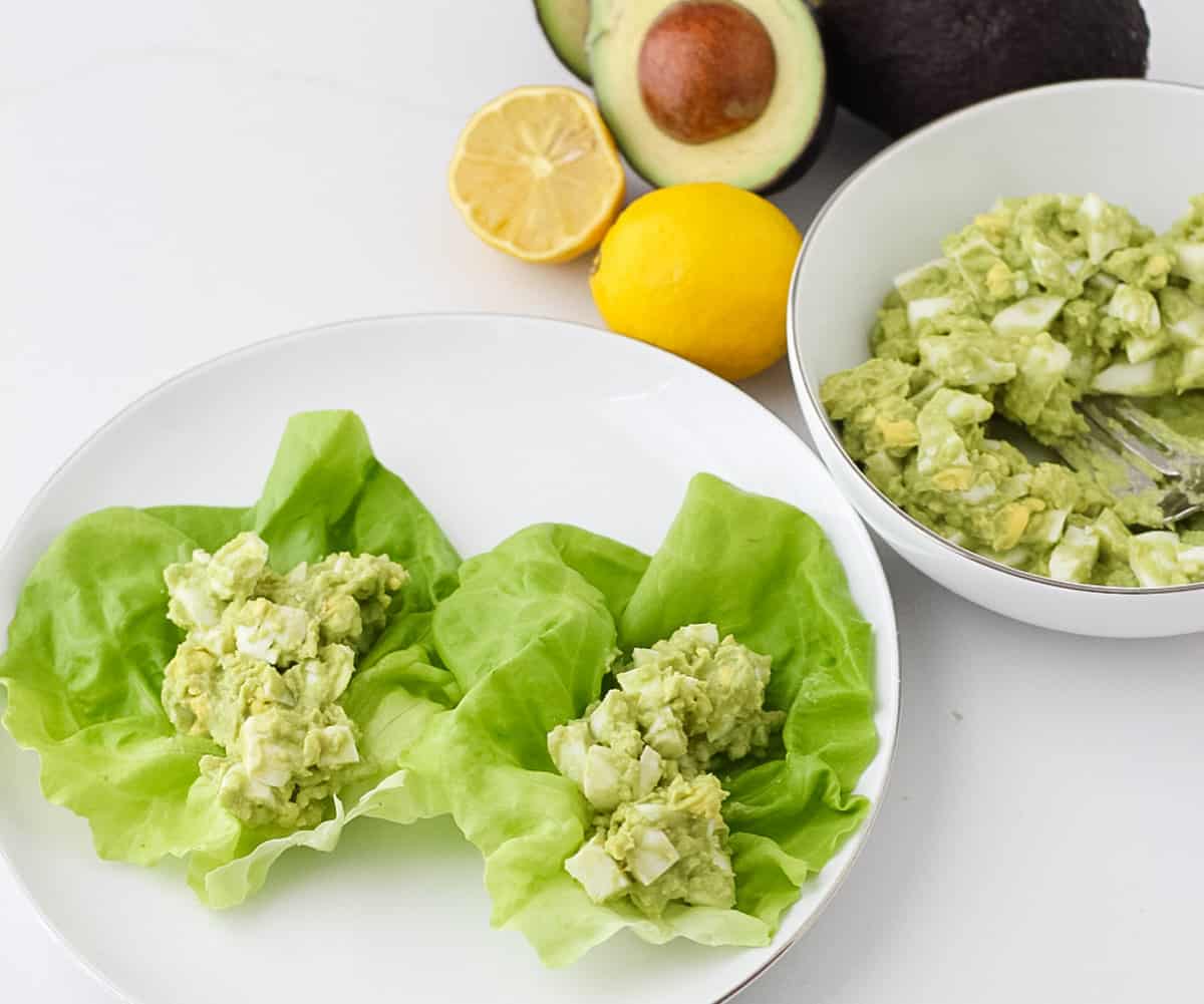 white plate with lettuce topped with egg salad made with avocado next to a bowl of avocado egg salad and fresh avocado and lemons.