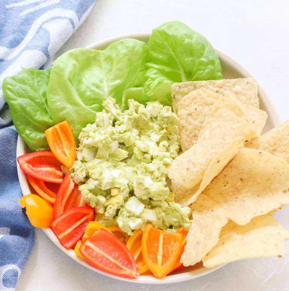 blue napkin next to a plate of avocado egg salad, tortilla chips, lettuce, and baby bell peppers.