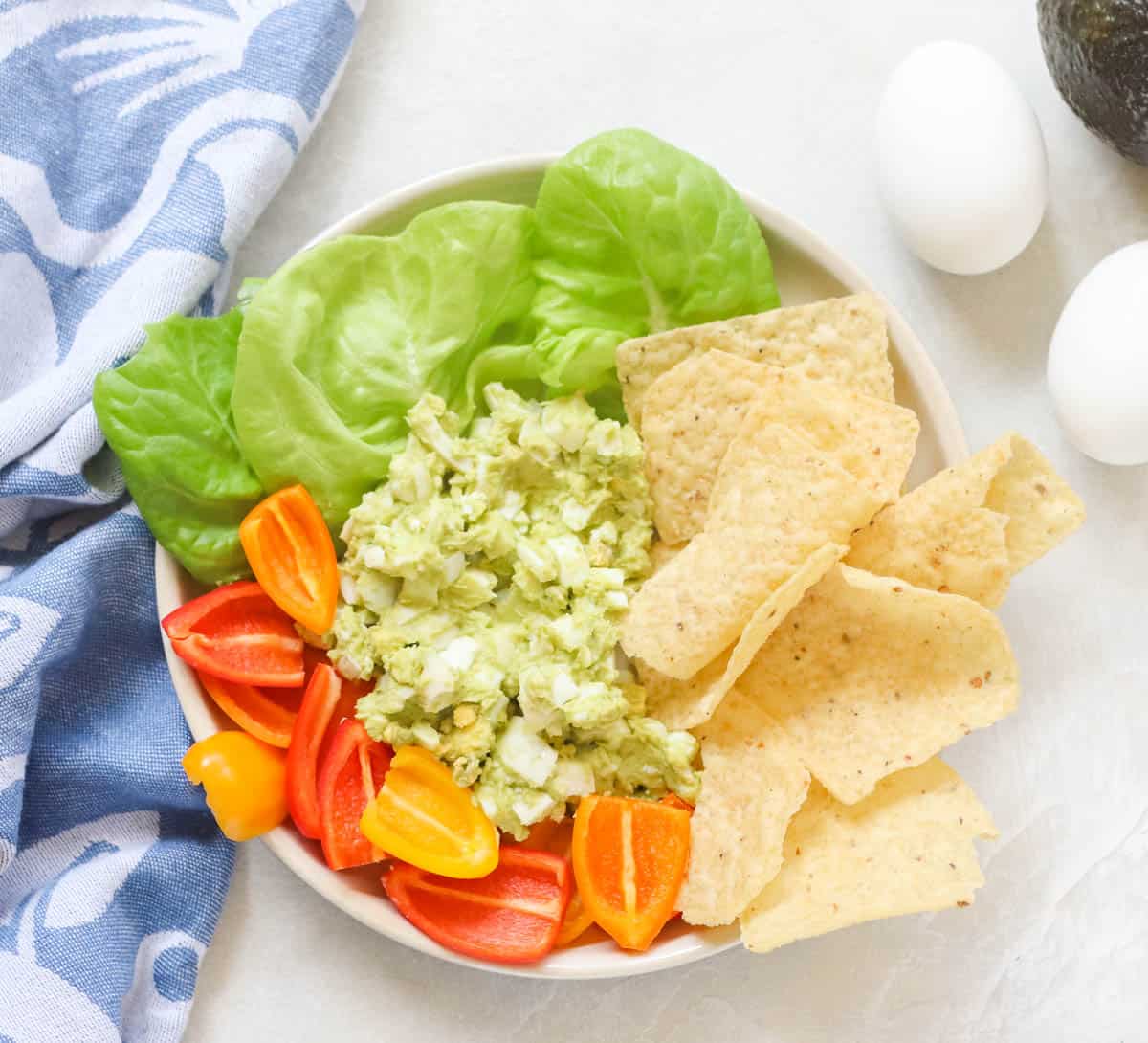 plate of lettuce, mini bell peppers, tortilla chips, and avocado egg salad next to a blue napkin and hardboiled eggs and avocado.