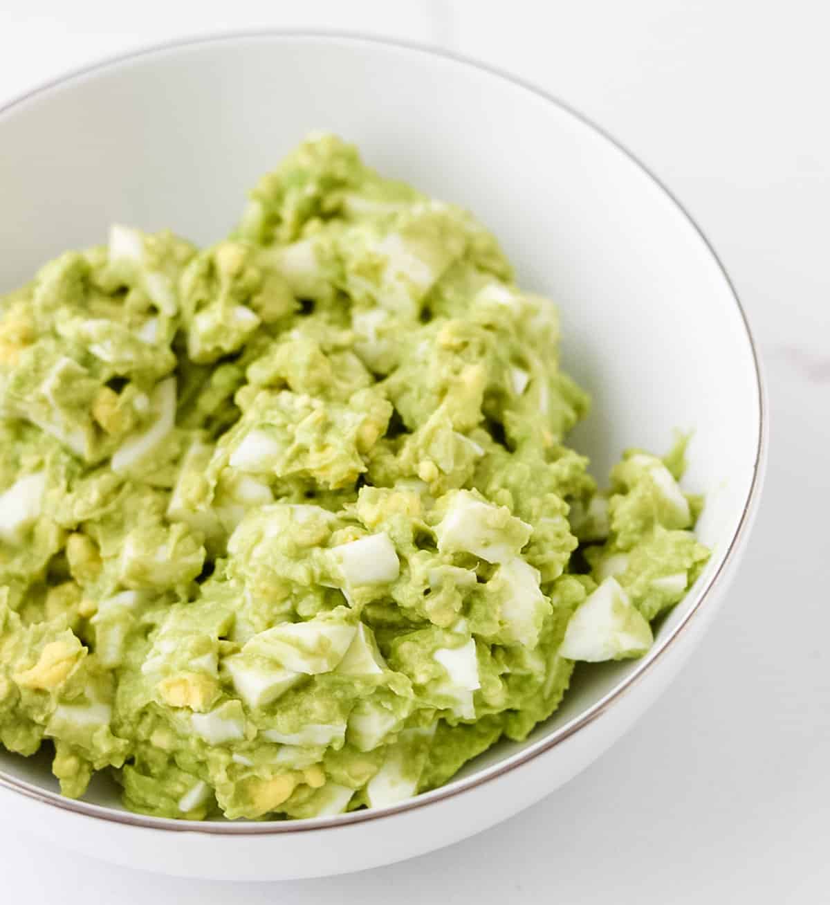 a white bowl filled with avocado egg salad on white countertop.