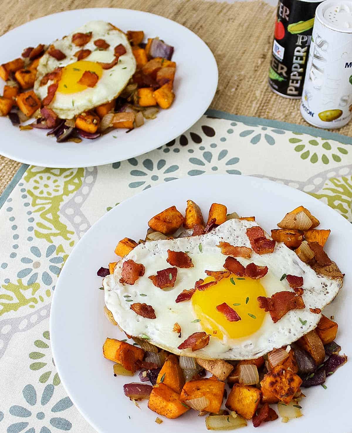 white plates with sweet potato bacon hash and fried egg next to salt and pepper on a flower placemat.