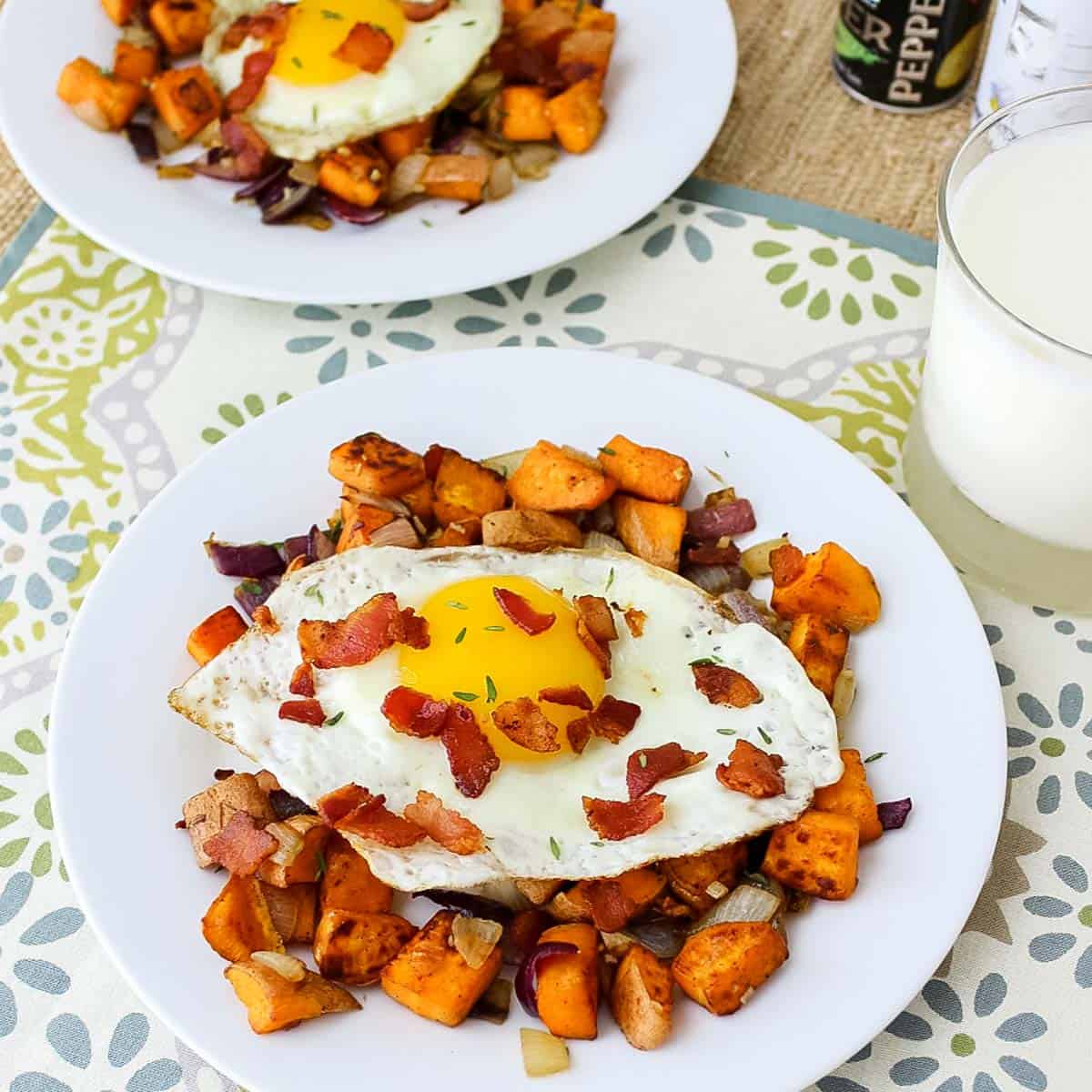 two white plates with sweet potato hash topped with an egg and bacon crumbles next to a glass of milk.