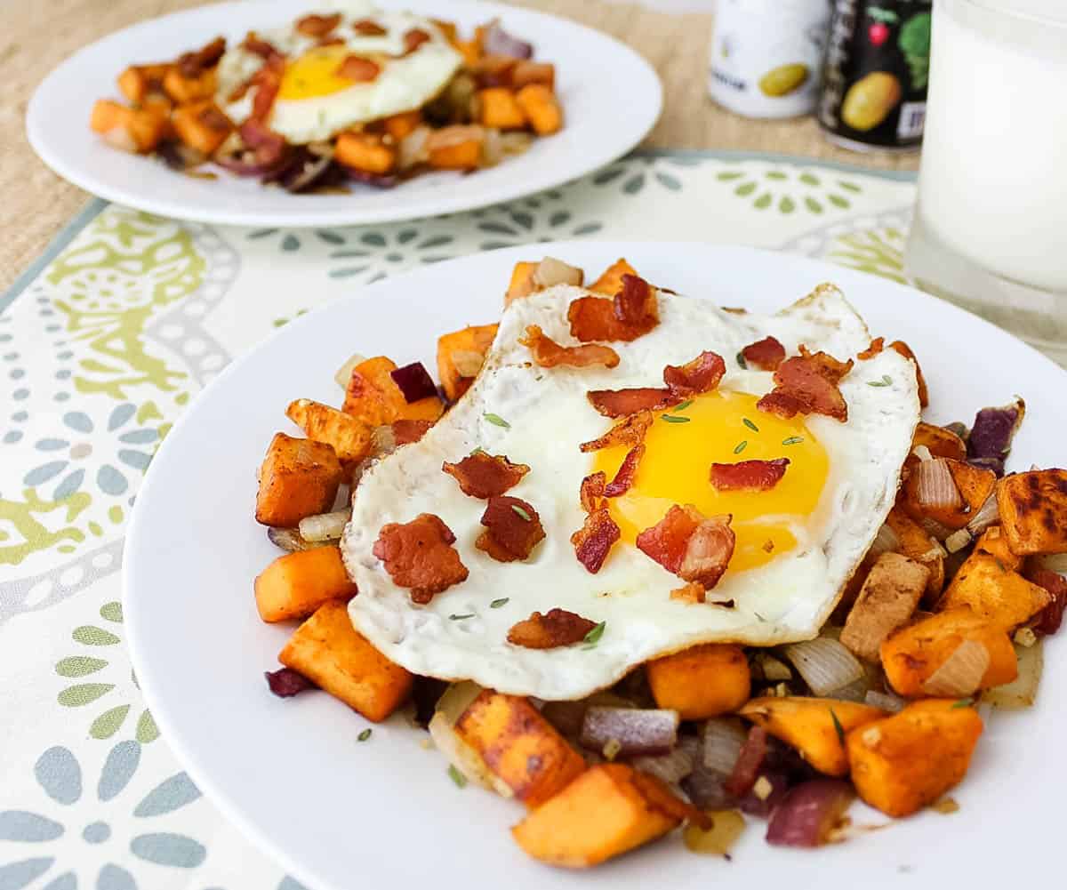 flower placemat with two plates of eggs and bacon topping sweet potato hash next to salt and pepper shakers and a glass of milk.