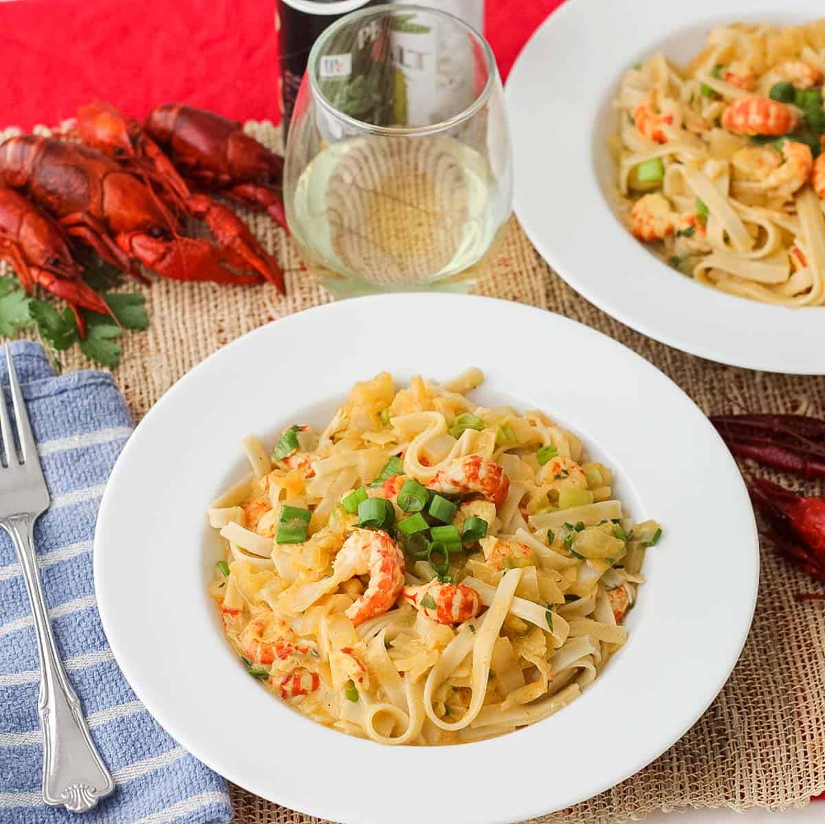 flat white bowls filled with fettuccine topped with crawfish Monica sauce, next to a blue napkin and fork, surrounded by a glass of white wine plus fresh boiled crawfish.