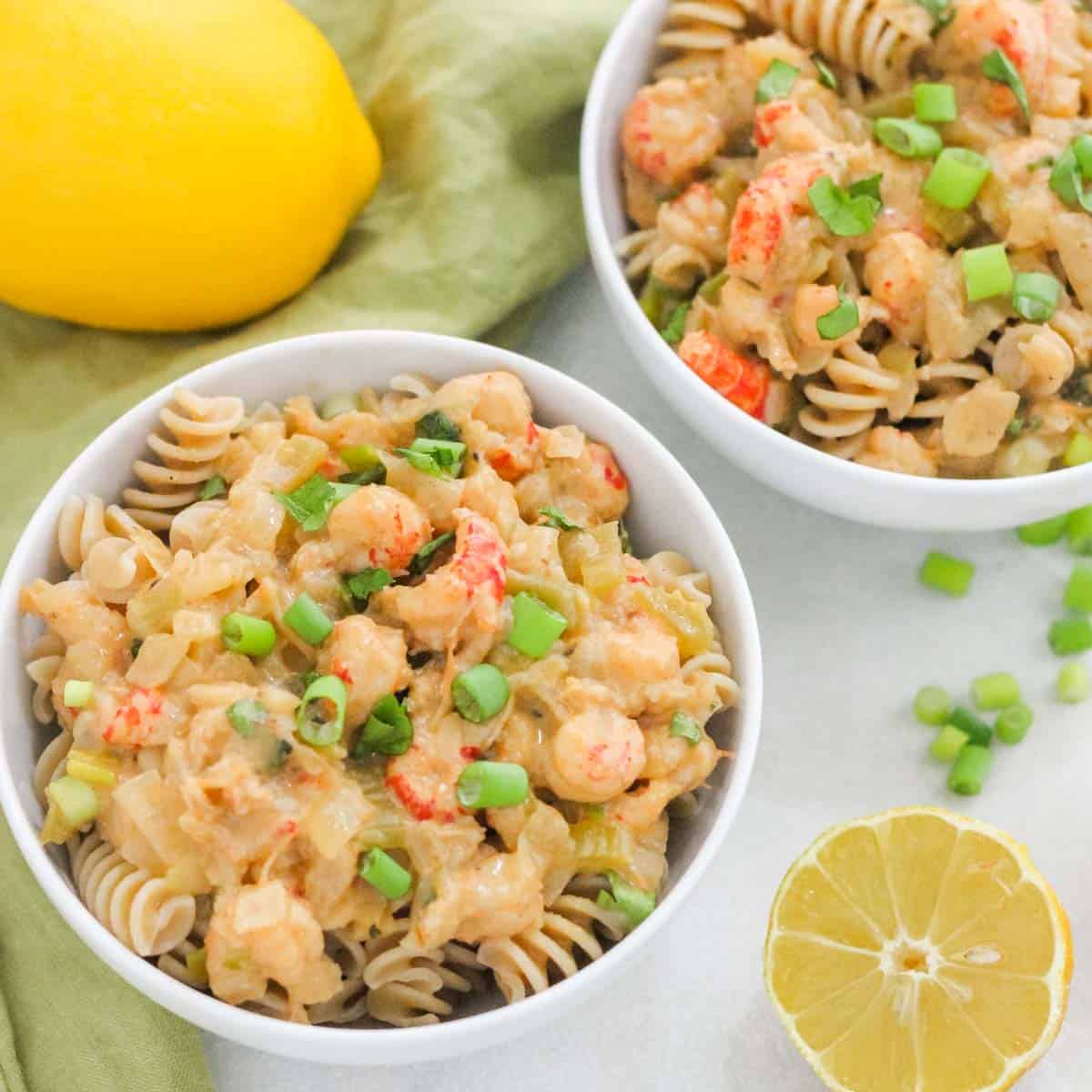 two bowls of rotini topped with crawfish Monica sauce next to lemons, green onions, and a green napkin.