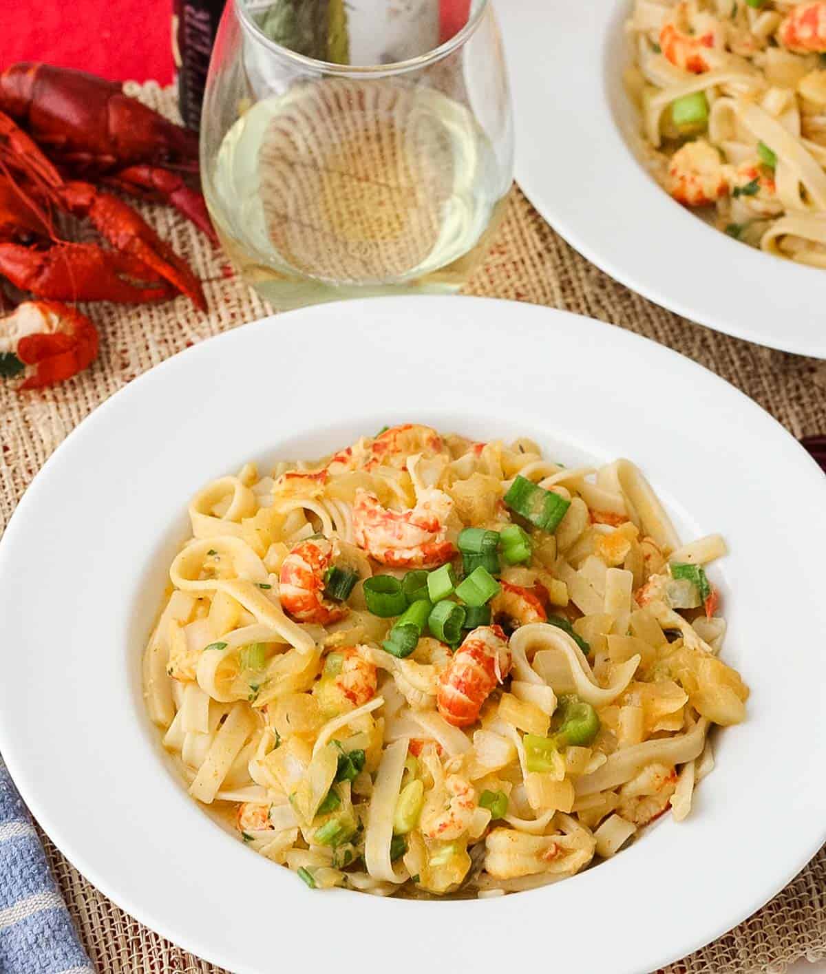 fettuccine topped with Crawfish Monica sauce and green onions in a white bowl next to a glass of white wine and freshly boiled crawfish.