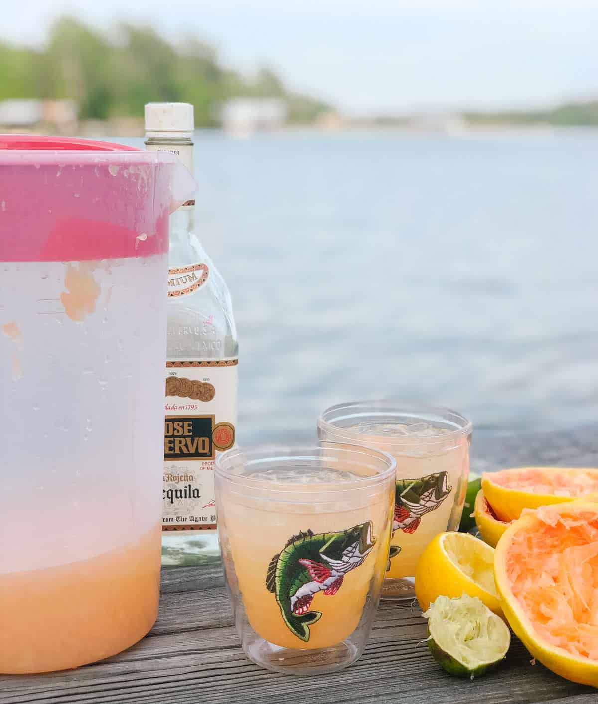 pitcher of margaritas, bottle of tequila, two cups with fish on it filled with grapefruit margaritas, juiced fruit all on a wooden table overlooking the water.