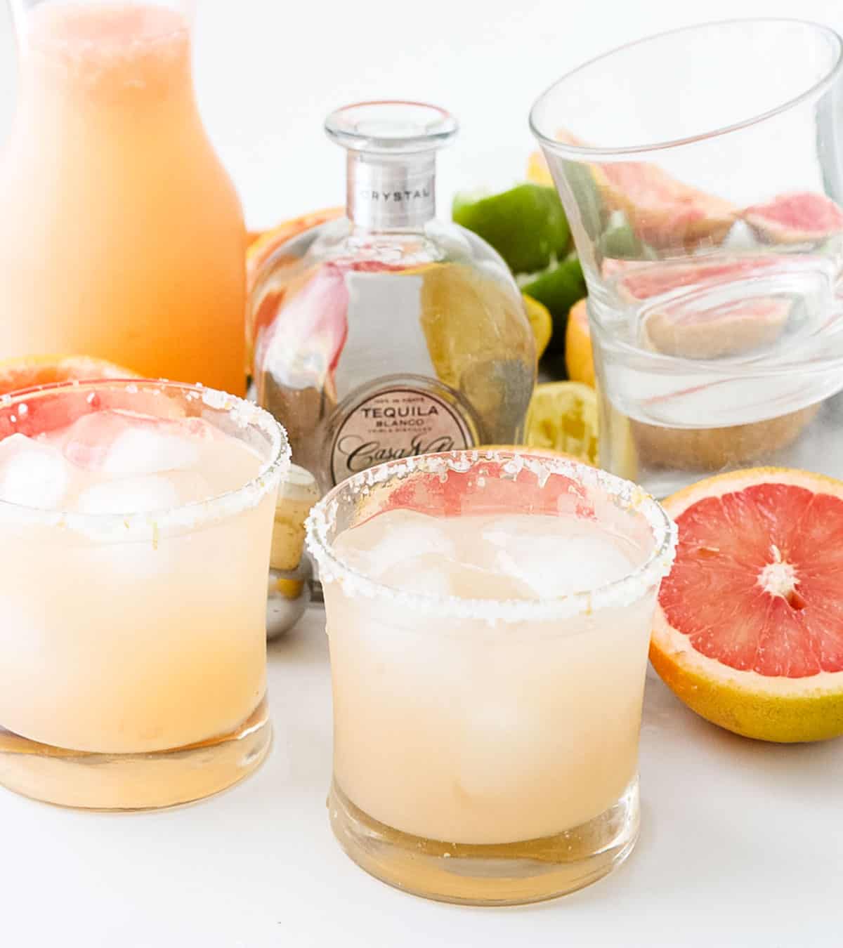 two salt-rimmed glasses with pink margaritas, red grapefruit, tequila, pitcher of grapefruit margarita, empty glasses.