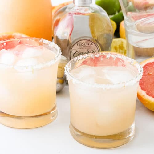 two glasses of pink margaritas next to a tequila bottle, red grapefruits, and a pitcher of grapefruit margaritas.