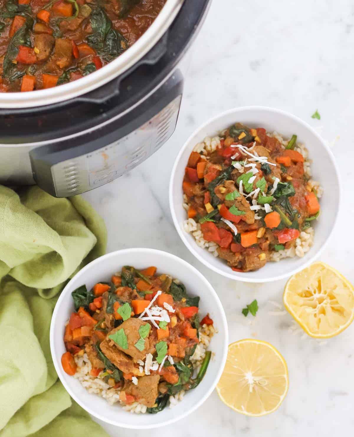 two bowls of lamb curry with mint and coconut flakes next to an instant pot, a green napkin, and cut lemons.