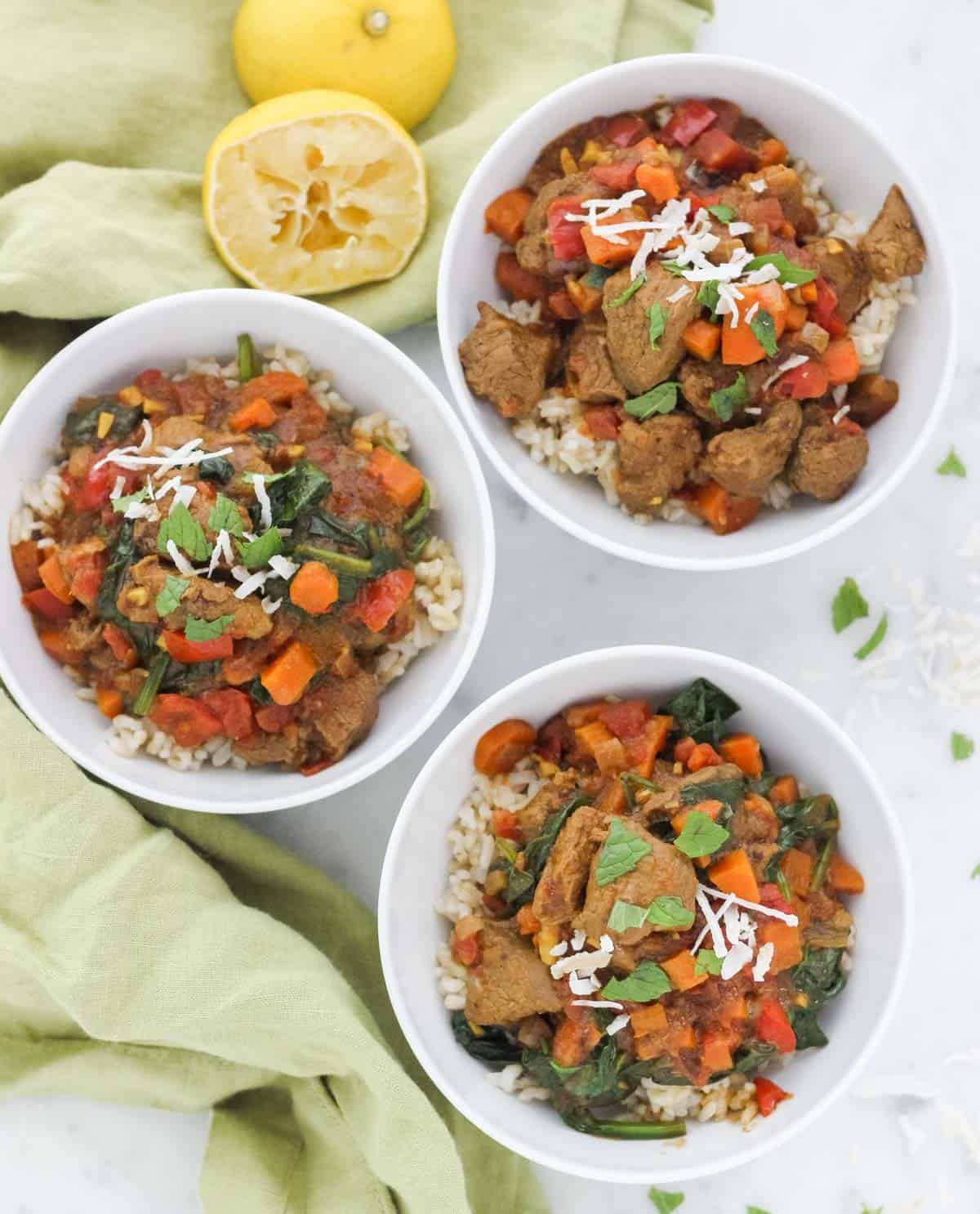 green napkin next to three bowls with lamb curry over brown rice sprinkled with mint and coconut flakes.