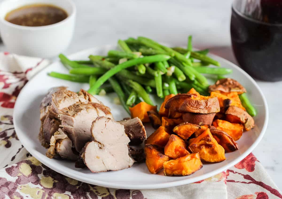 flower placemat with a white plate filled with pork tenderloin, roasted sweet potatoes, and green beans next to a glass of red wine.