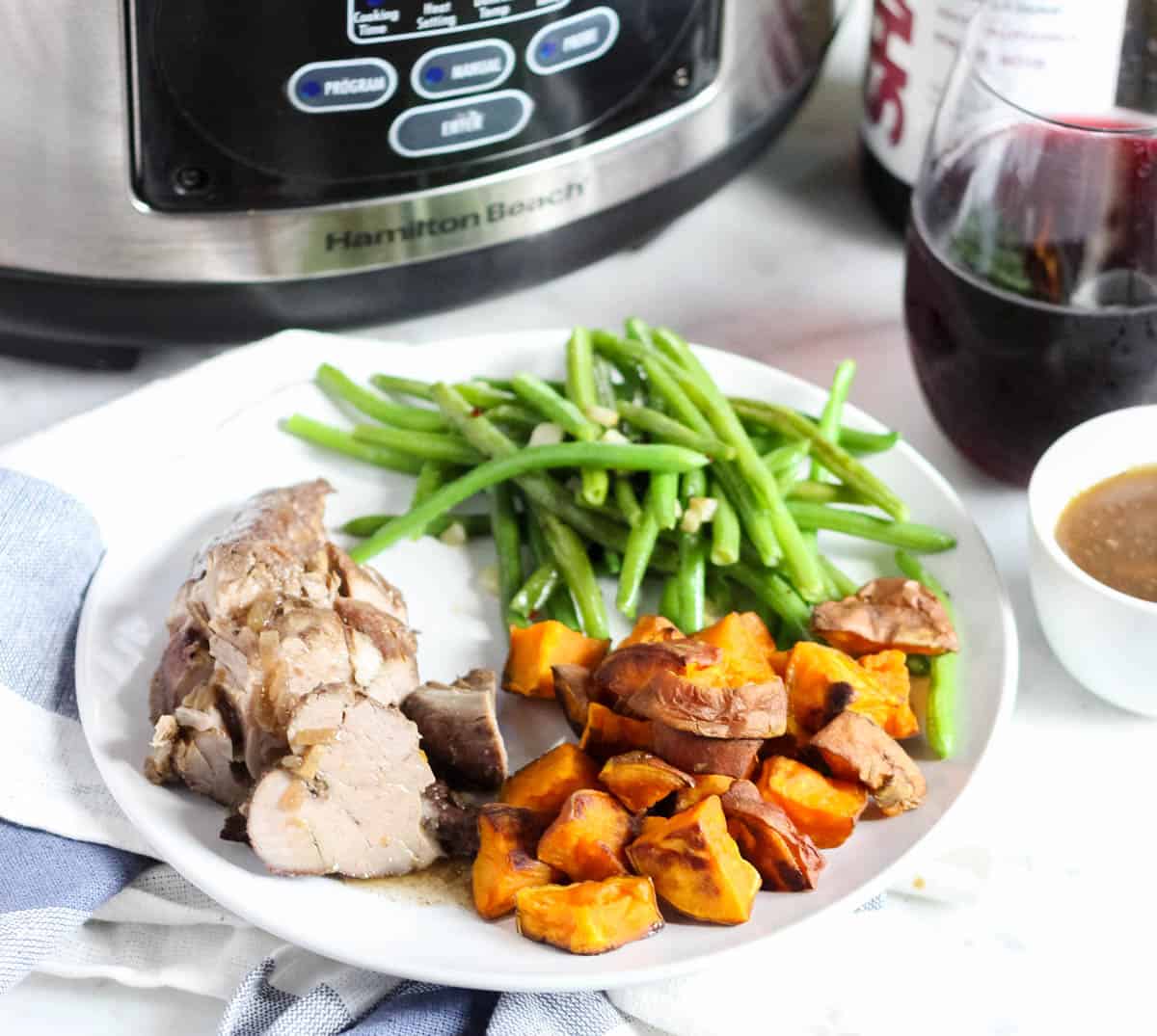 slow cooker, bottle of wine, glass of wine, and a bowl of gravy next to a plate of pork tenderloin, green beans, and roasted sweet potatoes.