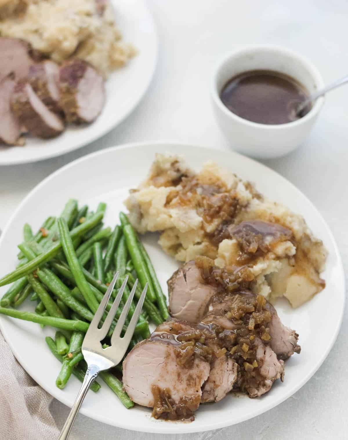 fork on a plate with green beans, pork tenderloin, and mashed potatoes next to a bowl of gravy.