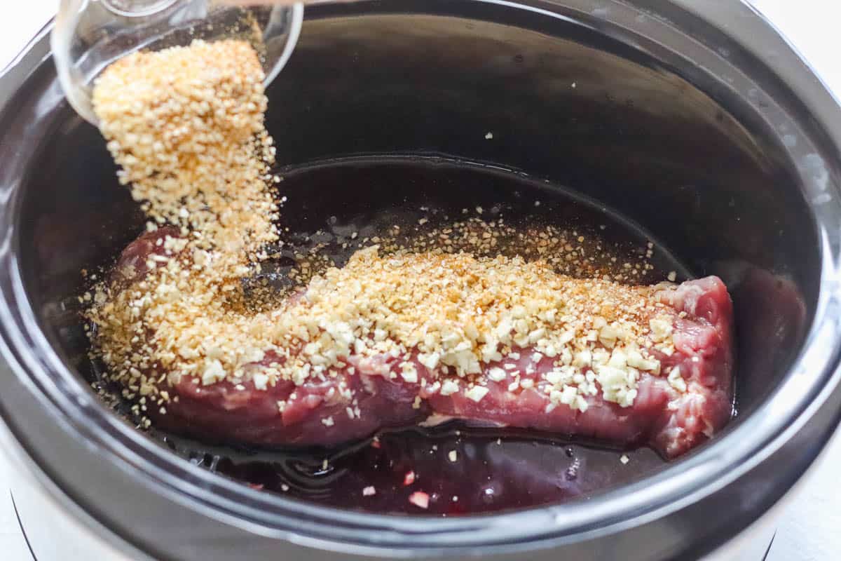 spices in a bowl being sprinkled over pork tenderloin in a crock pot.