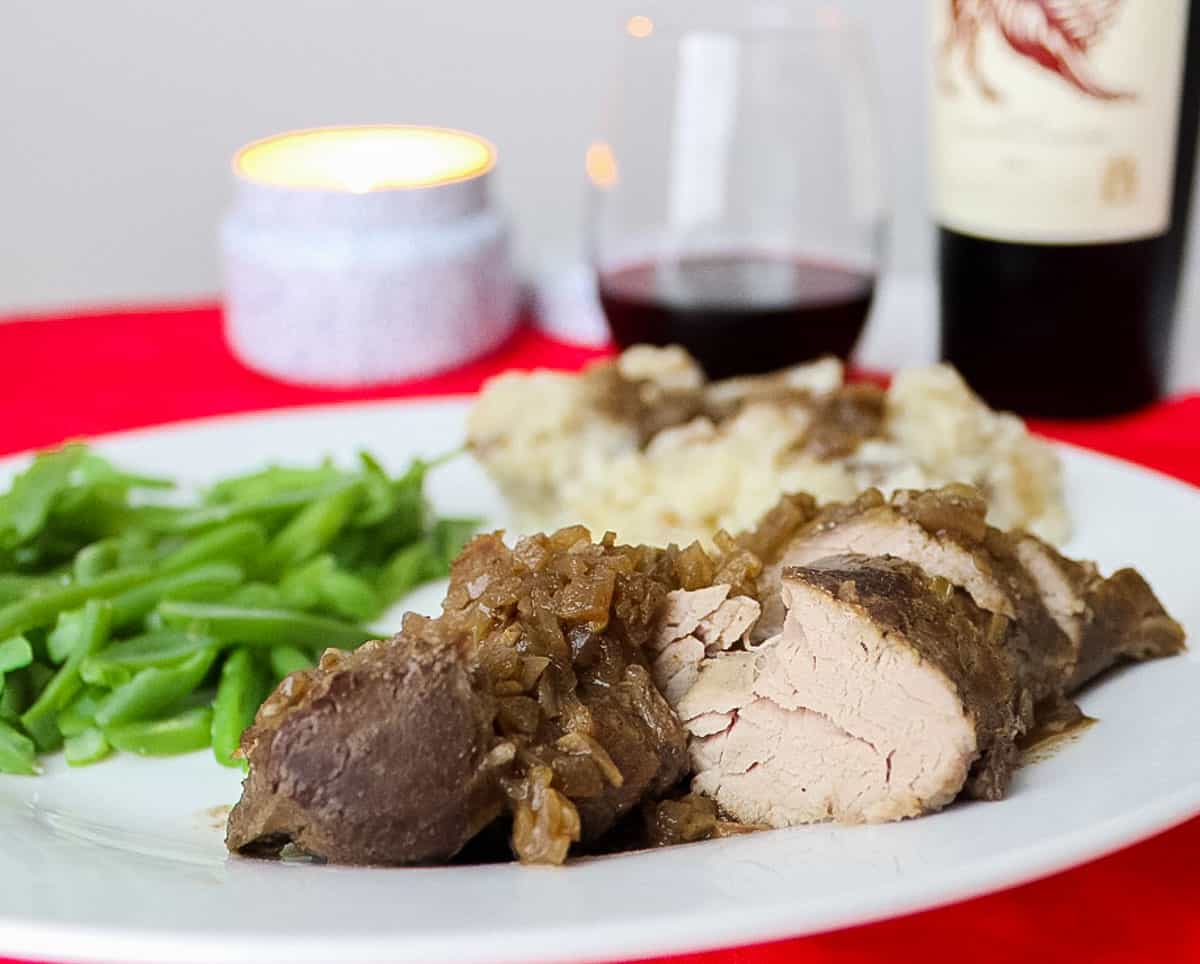 red placemat with a candle, bottle of red wine, glass of wine, and pork tenderloin covered in gravy next to mashed potatoes and green beans on a white plate.