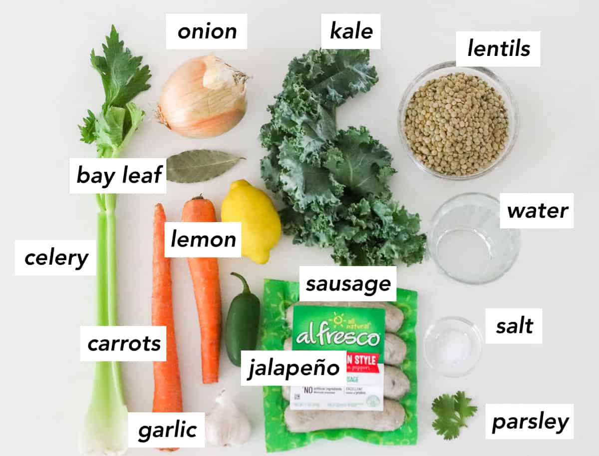 celery stick, two carrots, garlic, package of sausage, parsley, bowl of salt, cup of water, bowl of lentils, kale, lemon, jalapeno, bay leaf, onion.