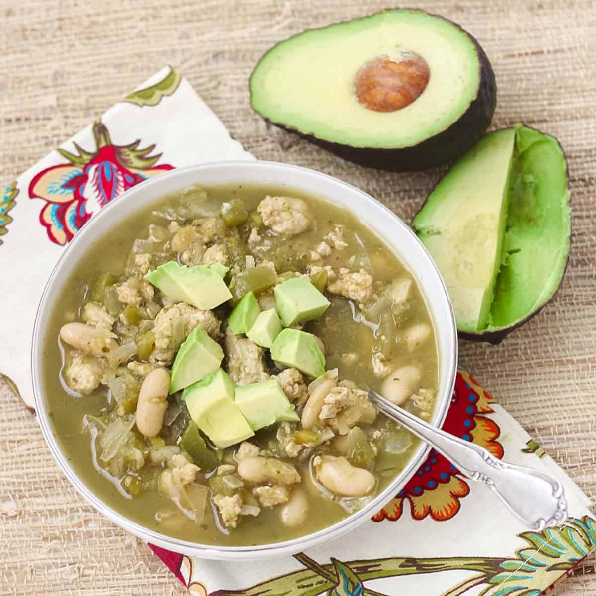 silver spoon in a bowl of chili topped with fresh avocado on a flower napkin and bamboo placemat.