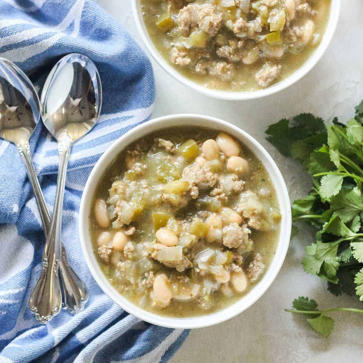 two silver spoons with two bowls of white turkey chili next to fresh cilantro.