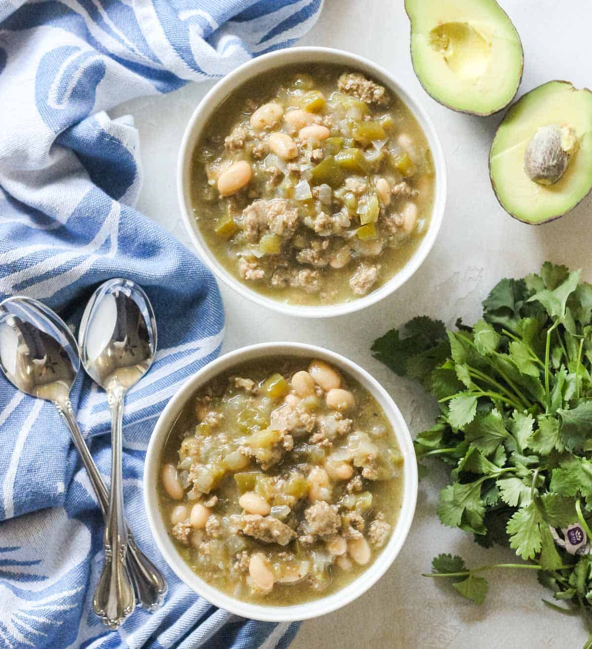 blue napkin with silver spoons next to two white bowls filled with white bean turkey chili next to fresh cilantro and avocados.