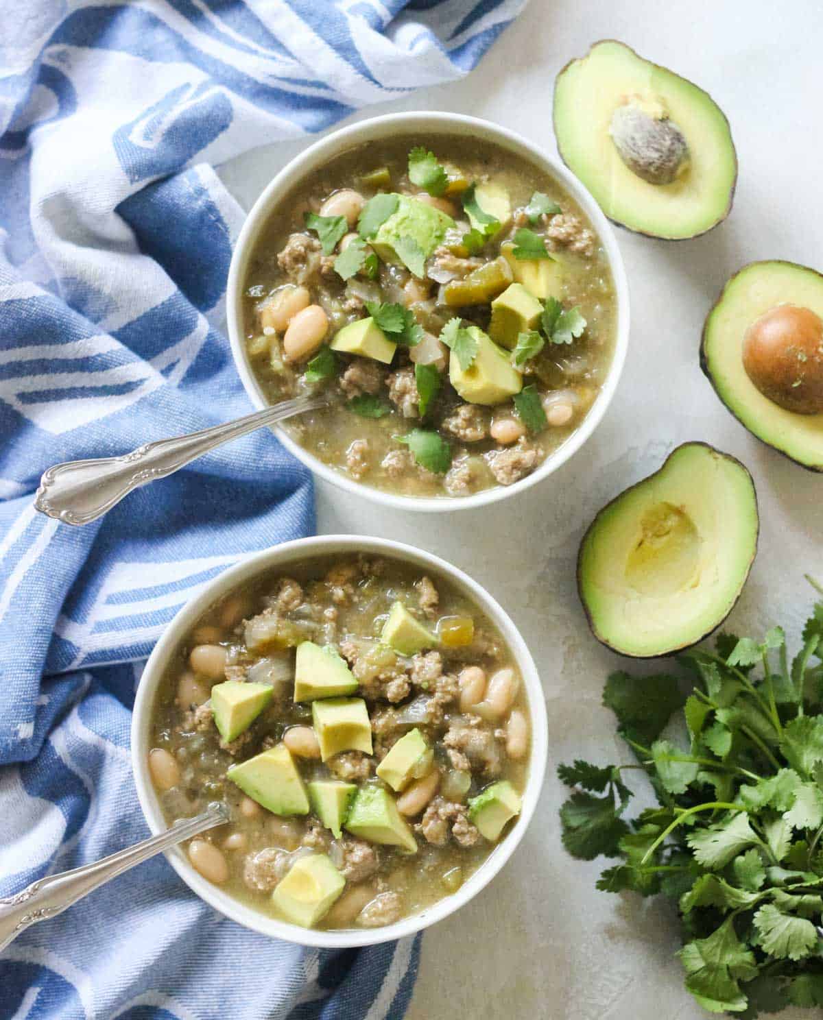 two bowls of chili topped with cilantro and avocado with silver spoons next to fresh cilantro and avocado.