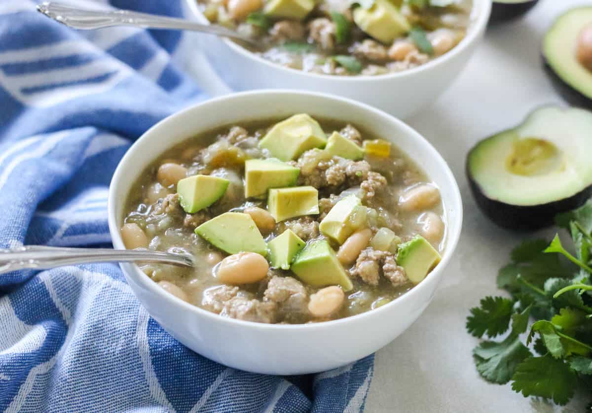 white bean and turkey chili in a white bowl with a silver spoon topped with fresh avocado next to fresh cilantro and a blue napkin.