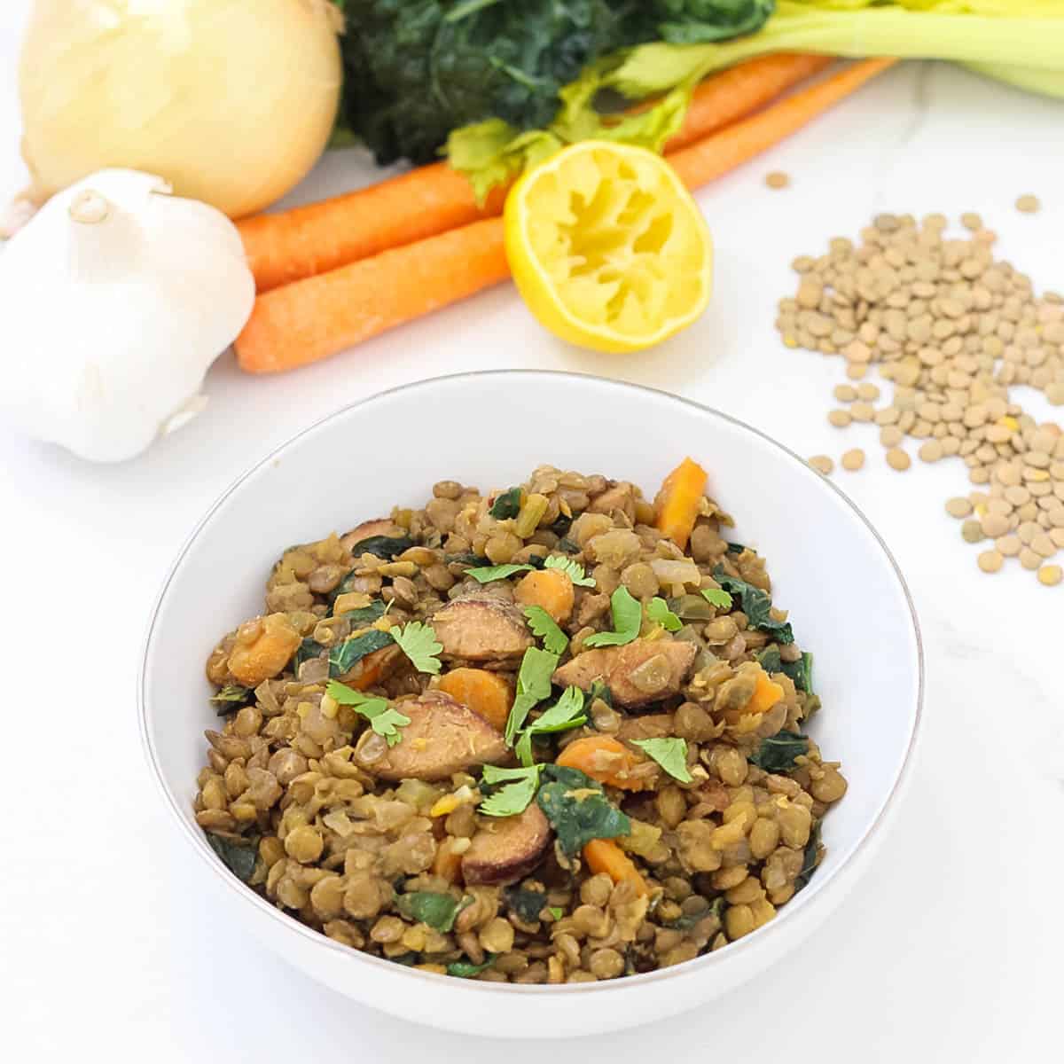 white bowl with sausage and lentils casserole topped with parsley surrounded by fresh garlic, lemon, carrots, celery, kale, and dried lentils.