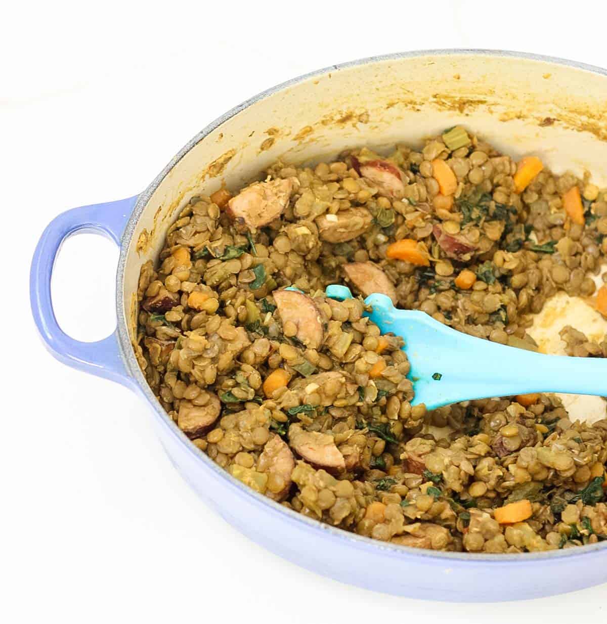 blue pot full of sausage and lentils with kale and veggies being stirred by a blue spoon.