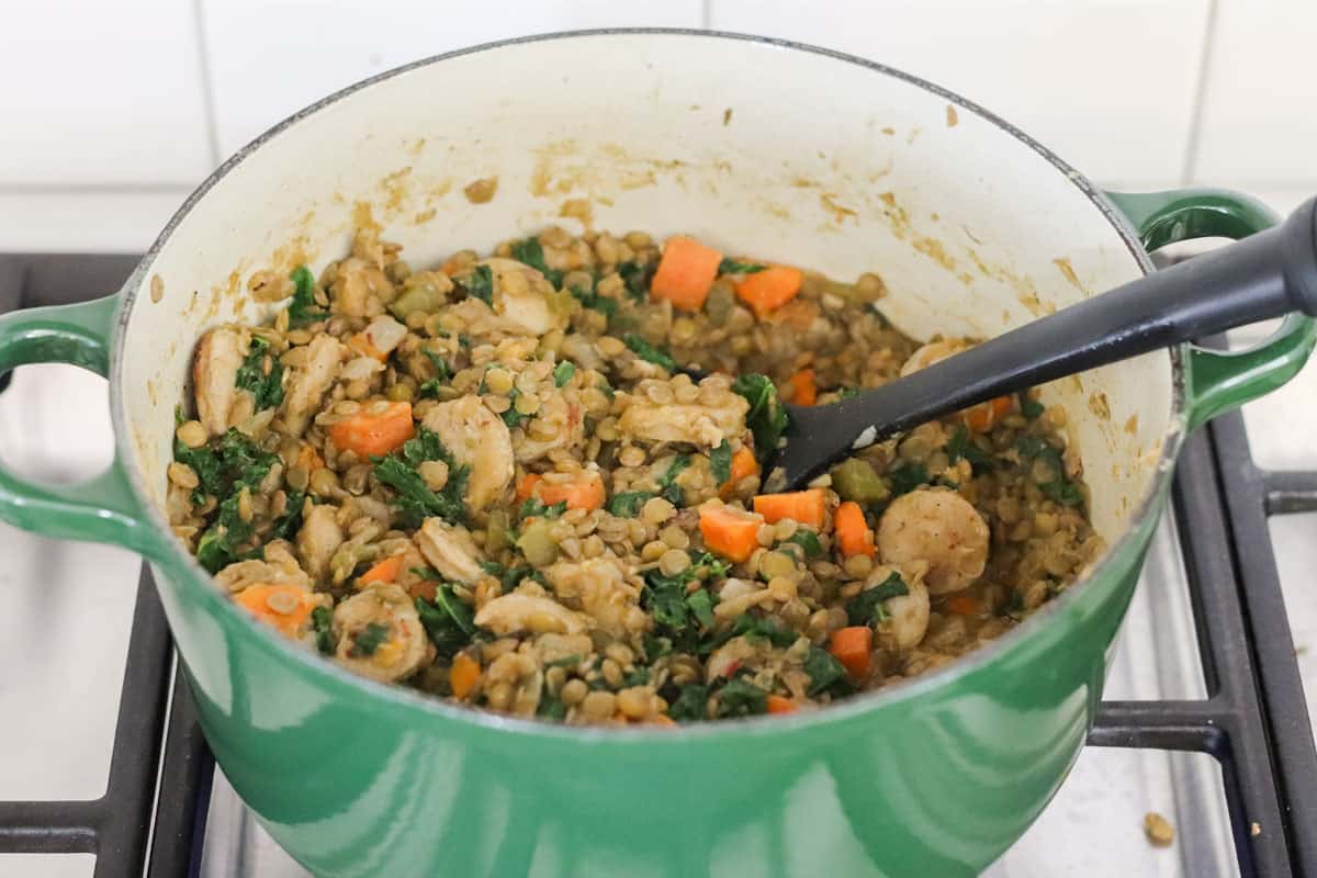 green pot with cooked lentils, sausage, kale, and carrots on a stovetop.
