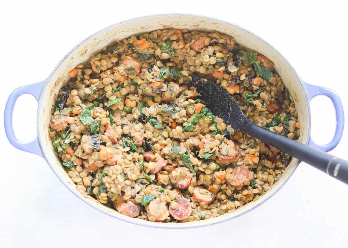 blue pot with cooked lentils, kale, and sausage being stirred with a black spoon.