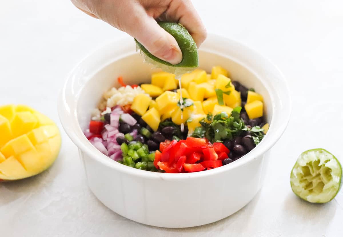 hand squeezing lime juice into a bowl of mango, black beans, onion, peppers, and garlic with a cut mango and lime nearby.