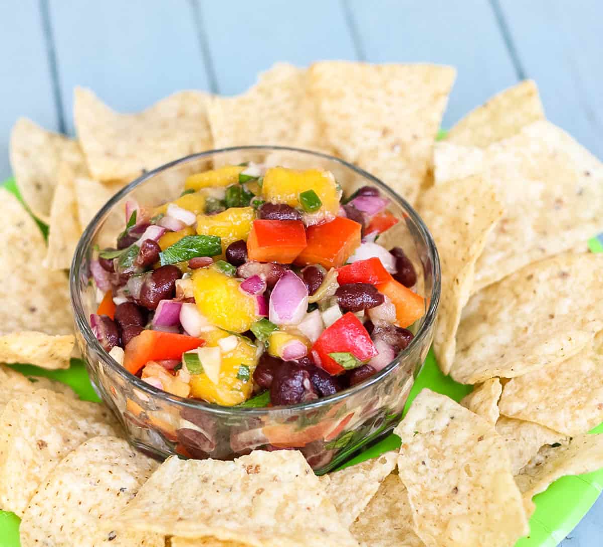 mango salsa with black beans in a clear bowl surrounded by tortilla chips on a green plate.