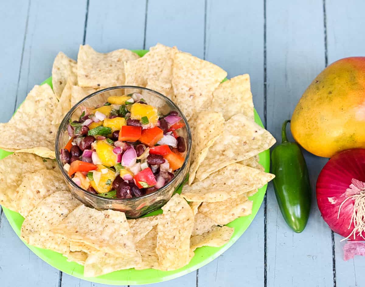 blue table with fresh mango, jalapeno, and red onion, plus a green plate with tortilla chips and mango salsa.