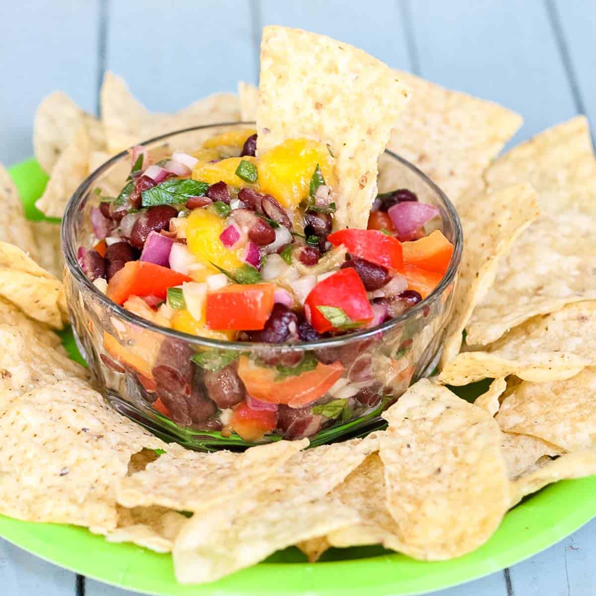 clear bowl with Mango and Black Bean Salsa on a green plate surrounded by tortilla chips on a blue table.