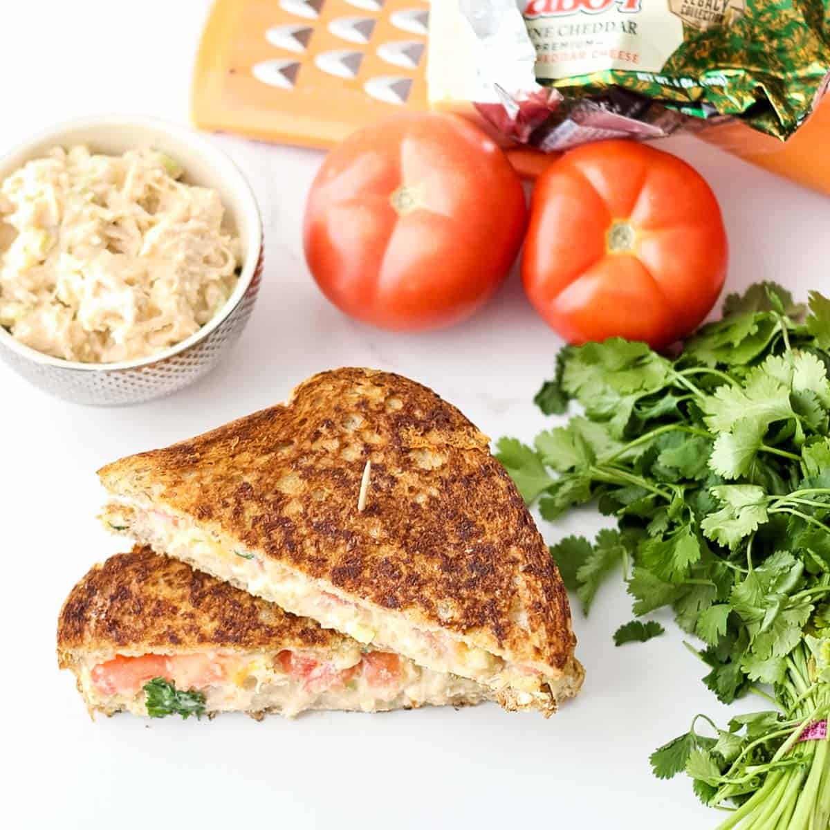 chicken salad grilled cheese on white counter next to fresh parsley, a bowl of chicken salad, fresh tomatoes, and block of cheddar cheese.