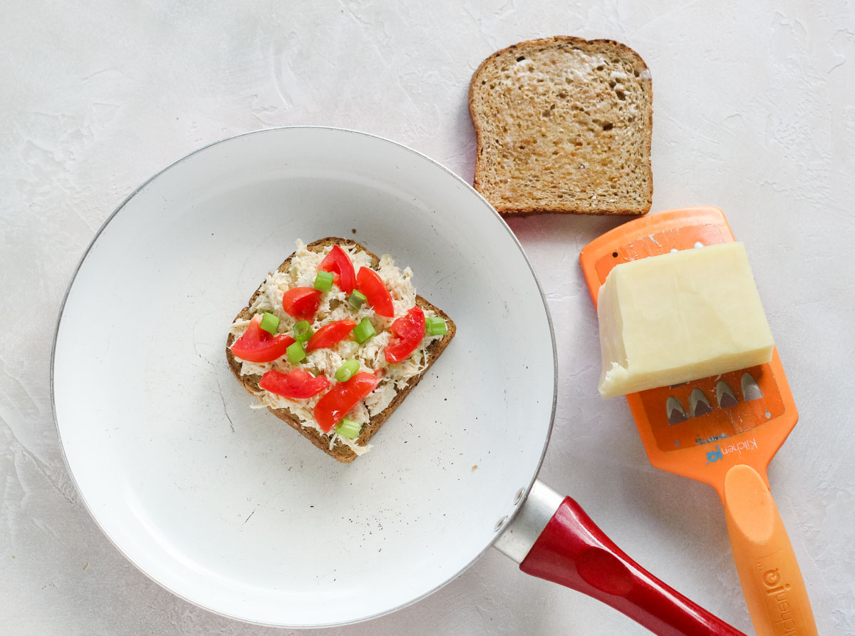white skillet with open face chicken salad sandwich topped with tomato and green onion next to a cheese grater with cheddar cheese and a slice of buttered bread.