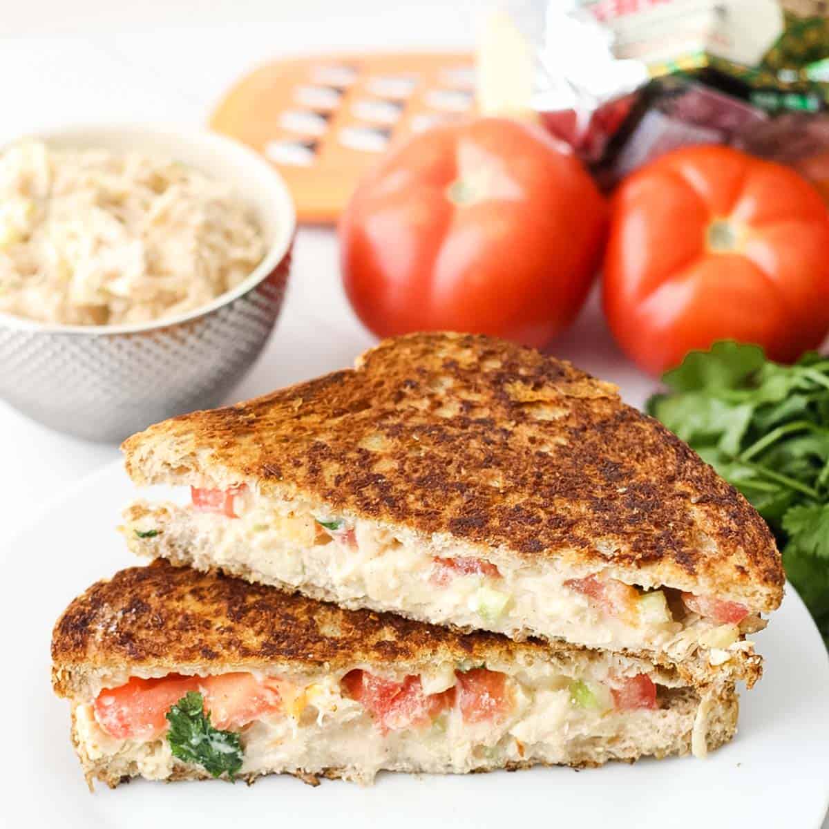 stacked chicken salad melt sandwich surrounded by fresh tomatoes, parsley, and a bowl of chicken salad.
