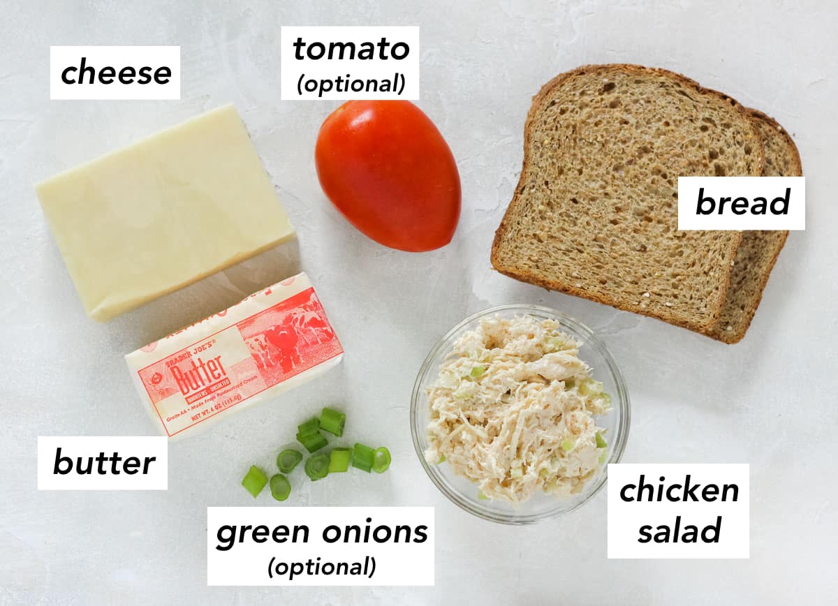 block of white cheddar cheese, stick of butter, chopped green onion, bowl of chicken salad, two slices of whole grain bread, and a roma tomato on a white counter with text overlay describing ingredients.