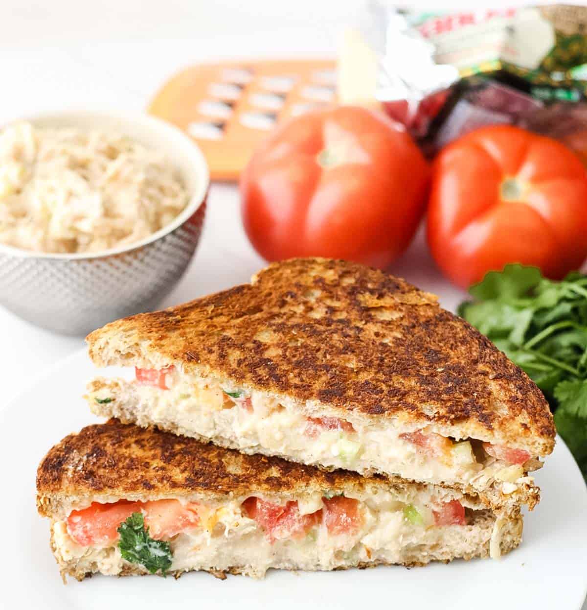 stacked chicken salad melt sandwich surrounded by tomatoes, parsley, and a bowl of chicken salad.